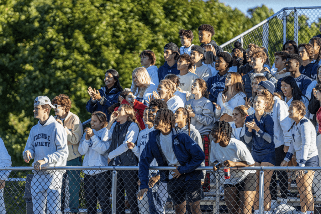 A large group of Cheshire Academy students on the bleachers anxiously cheer on their friends at a sports game.