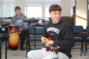 student playing guitar in open class room, student in far distance also playing an instrument,
