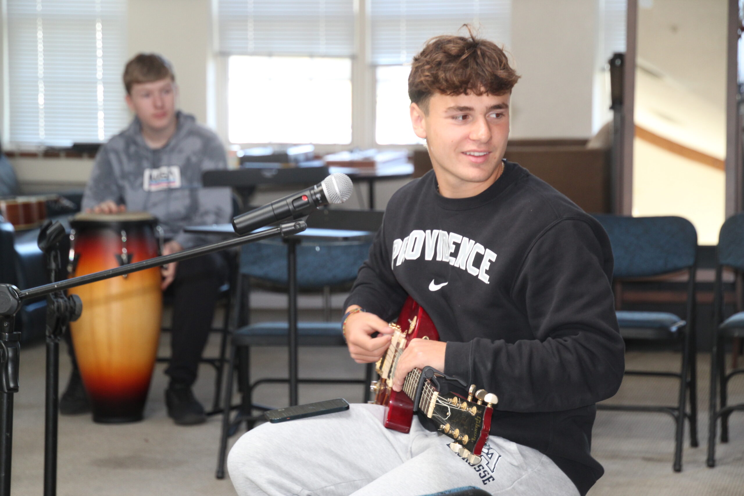 student playing guitar in open class room, student in far distance also playing an instrument,